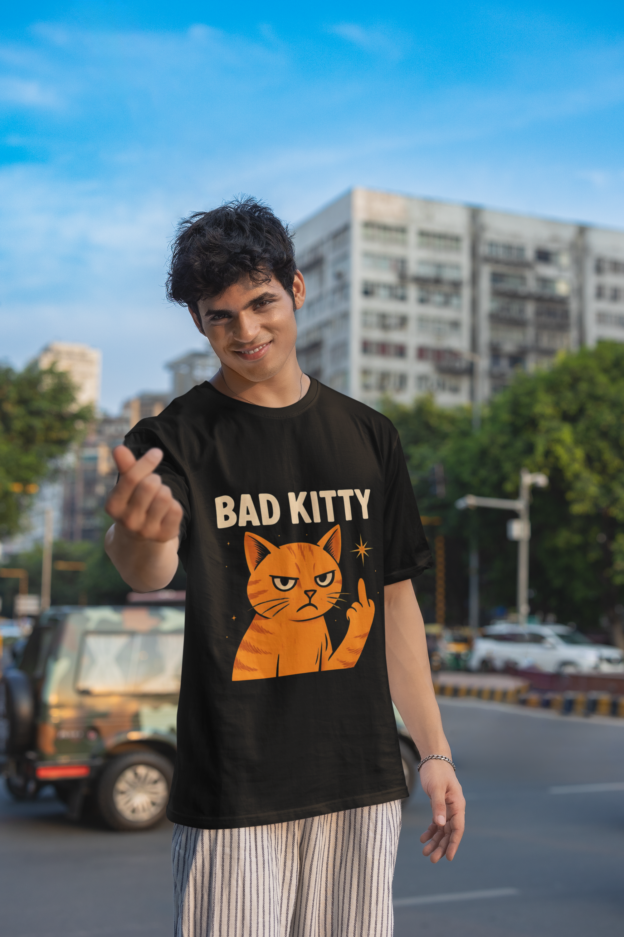 A man wearing a black "Bad Kitty" graphic t-shirt with an orange cat giving a middle finger, looking upwards with a serious expression. He is standing outdoors with a white building and a street in the background.