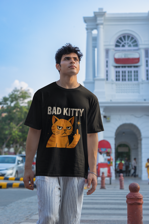 A man wearing a black "Bad Kitty" graphic t-shirt with an orange cat giving a middle finger, looking upwards with a serious expression. He is standing outdoors with a white building and a street in the background.