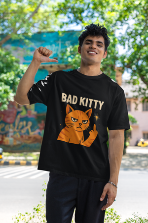 A man wearing a black "Bad Kitty" graphic t-shirt with an orange cat giving a middle finger, looking upwards with a serious expression. He is standing outdoors with a white building and a street in the background.