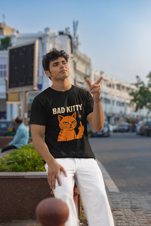 A man wearing a black "Bad Kitty" graphic t-shirt with an orange cat giving a middle finger, looking upwards with a serious expression. He is standing outdoors with a white building and a street in the background.