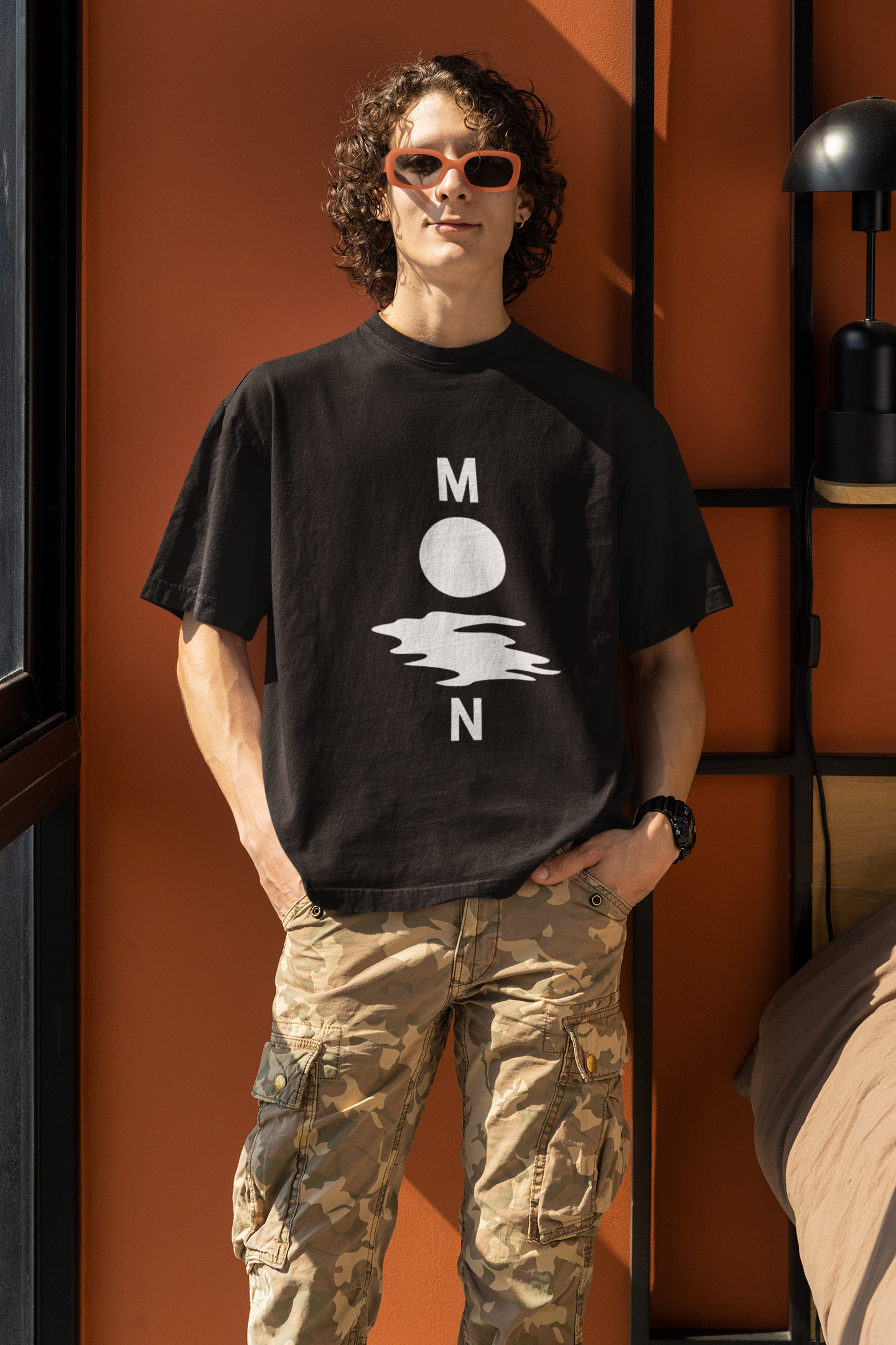 A young man with curly hair and sunglasses wearing a black oversized t-shirt with a white minimalist graphic depicting the letters "M" and "N" flanking a circle and a wavy line, spelling "MOON." He is wearing camouflage cargo pants and standing against an orange wall indoors.