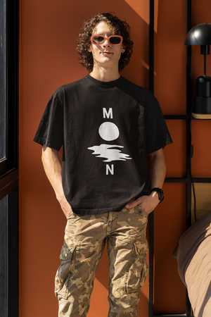 A young man with curly hair and sunglasses wearing a black oversized t-shirt with a white minimalist graphic depicting the letters "M" and "N" flanking a circle and a wavy line, spelling "MOON." He is wearing camouflage cargo pants and standing against an orange wall indoors.