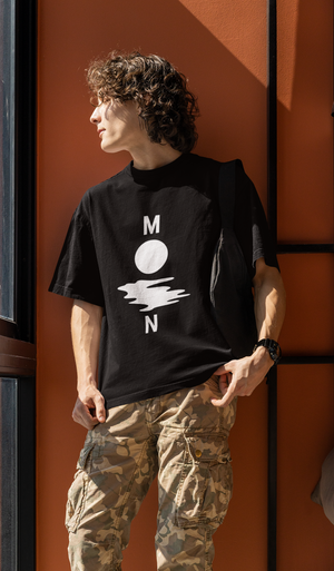 A young man with curly hair and sunglasses wearing a black oversized t-shirt with a white minimalist graphic depicting the letters "M" and "N" flanking a circle and a wavy line, spelling "MOON." He is wearing camouflage cargo pants and standing against an orange wall indoors.