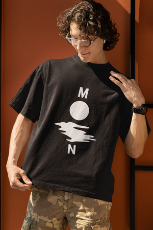 A young man with curly hair and sunglasses wearing a black oversized t-shirt with a white minimalist graphic depicting the letters "M" and "N" flanking a circle and a wavy line, spelling "MOON." He is wearing camouflage cargo pants and standing against an orange wall indoors.