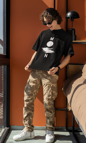 A young man with curly hair and sunglasses wearing a black oversized t-shirt with a white minimalist graphic depicting the letters "M" and "N" flanking a circle and a wavy line, spelling "MOON." He is wearing camouflage cargo pants and standing against an orange wall indoors.