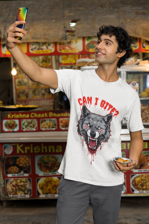 A smiling man wearing a white t-shirt with a fierce grey wolf graphic on the front, featuring red eyes, blood drips, and the text "CAN I BITE?". He is holding a brown cup and standing in an outdoor setting decorated with colorful tassels.