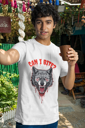 A smiling man wearing a white t-shirt with a fierce grey wolf graphic on the front, featuring red eyes, blood drips, and the text "CAN I BITE?". He is holding a brown cup and standing in an outdoor setting decorated with colorful tassels.