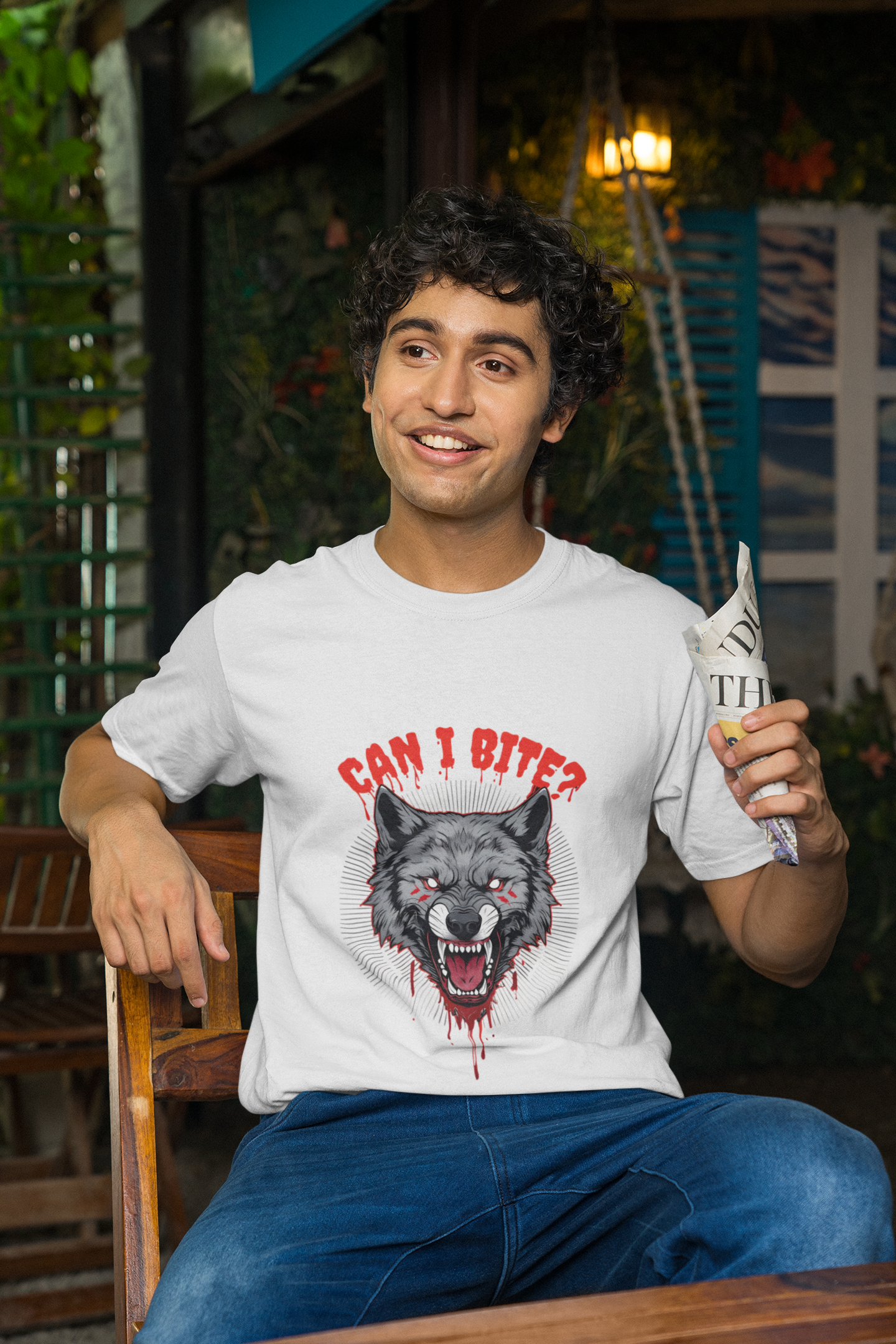 A smiling man wearing a white t-shirt with a fierce grey wolf graphic on the front, featuring red eyes, blood drips, and the text "CAN I BITE?". He is holding a brown cup and standing in an outdoor setting decorated with colorful tassels.