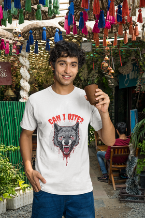 A smiling man wearing a white t-shirt with a fierce grey wolf graphic on the front, featuring red eyes, blood drips, and the text "CAN I BITE?". He is holding a brown cup and standing in an outdoor setting decorated with colorful tassels.
